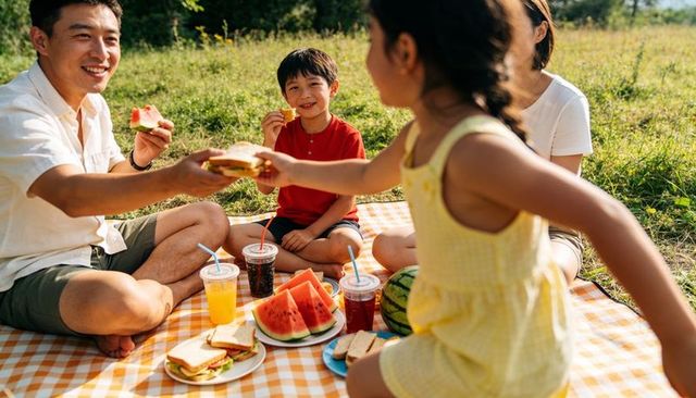 Family enjoying sunlit picnic in meadow sharing sandwiches, watermelon and laughter