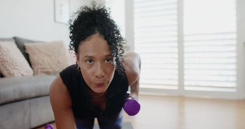 African american woman doing single-arm dumbbell row on mat in bright living room showing focus
