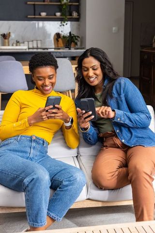 Diverse women sharing smartphones and laughing on sofa in modern open-plan living room