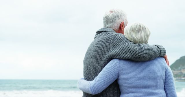 Senior couple with gray hair embracing while observing calm sea during winter. They wear cozy knit sweaters, highlighting their closeness and deep bond. Ideal for retirement themes, senior lifestyle topics, love and companionship stories, health and well-being content, and travel and leisure promotions targeted at the elderly.
