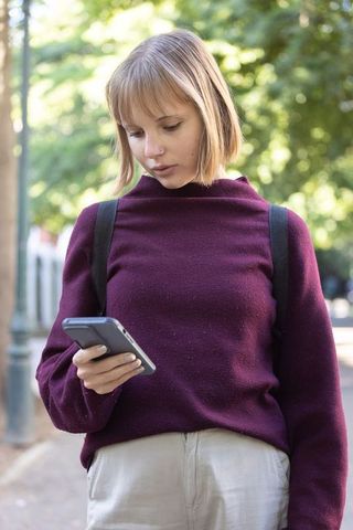Young woman checking smartphone on tree-lined sidewalk wearing maroon sweater and backpack