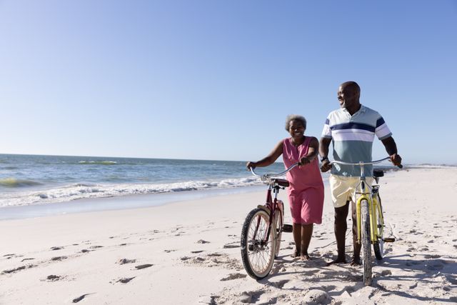 Senior African American couple walking with bicycles on a sandy beach under a clear blue sky. Ideal for concepts related to retirement, travel, healthy lifestyle, love, and enjoying nature. Perfect for use in advertisements, travel brochures, and articles about senior activities and vacations.