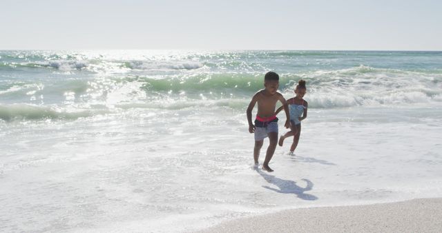 Children enjoying a sunny day at the beach, running along the shoreline with waves in the background. This image can be used for advertising family vacations, beach resorts, outdoor activities, or promoting a healthy, active lifestyle for children.