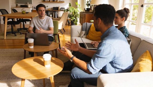 Sunlit casual team meeting in cozy living room coworking with laptops and takeaway coffee