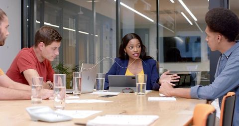 Woman Leading Team Meeting in Glass-Walled Conference Room Using Tablet and Laptop