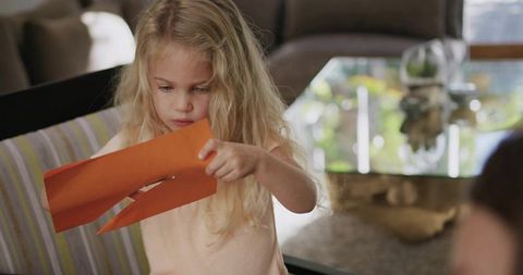 Blonde girl crafting with orange paper cutting strips, concentrating on hands-on learning