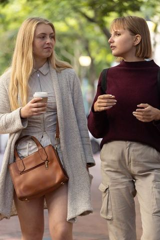 Young women walking and talking on tree-lined city sidewalk holding coffee and handbag