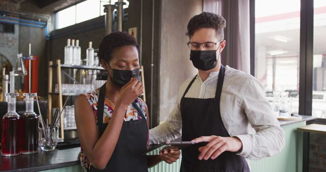 Portrait of diverse couple wearing face masks working at a bar, using tablet and smiling to camera. work at an independent bar business during coronavirus covid 19 pandemic.