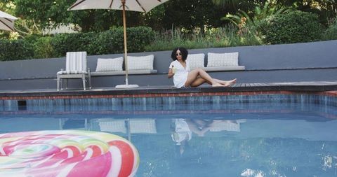 African american woman relaxing poolside in white coverup holding cocktail by lollipop float