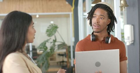 Diverse coworkers chatting in modern coworking space holding laptop and wearing headphones