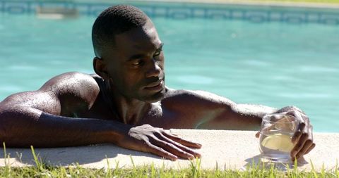 African american man relaxing at pool edge holding glass of cold beverage in sunshine
