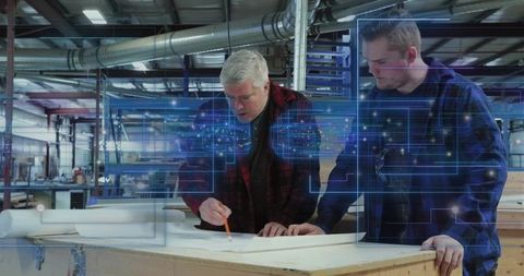 Senior craftsman mentoring young technician over woodworking blueprints with ar overlay in workshop