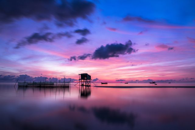 Silhouetted fishing hut stands on stilts against a stunning pink and purple sunset, reflecting over a calm ocean. Idle boats and scattered clouds decorate the horizon, creating a tranquil and serene atmosphere. Suitable for travel websites, inspirational posters, or promoting relaxation and mindfulness.