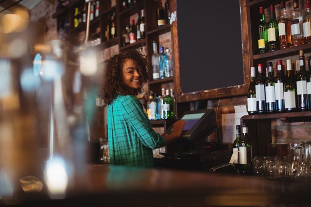 This image shows a cheerful female bartender with curly hair using a POS system at a bar counter. The background features shelves filled with various wine bottles, creating a lively and inviting atmosphere. This image is ideal for use in articles or advertisements related to hospitality, nightlife, bar management, customer service, and restaurant operations.