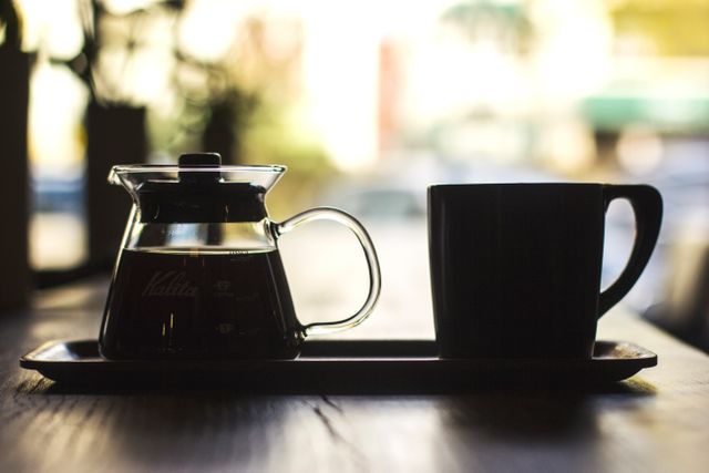 Brewed coffee in a glass pot and a mug on a wooden table in a cafe. Warm natural light coming through the window. Perfect for representing a peaceful and calm morning, cozy coffee shops, breakfast scenes, and moments of relaxation.