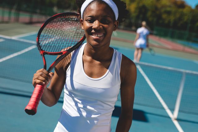 Young African American female tennis player smiling and holding a racket on her shoulder at an outdoor tennis court. Ideal for use in sports promotions, fitness campaigns, and advertisements for athletic wear. Perfect for illustrating themes of active lifestyle, competition, and recreation.