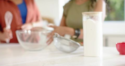 Diverse women baking together in bright kitchen pouring flour and whisking in bowl