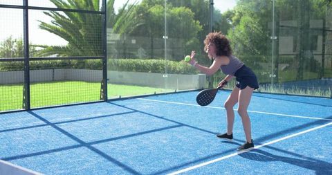 African american woman serving padel on blue court with sunlit glass walls and shadows