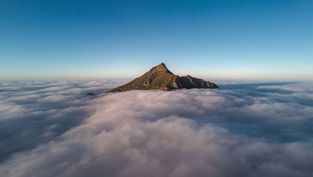 Rising alpine peak piercing sea of clouds at golden hour, aerial cloudscape serenity