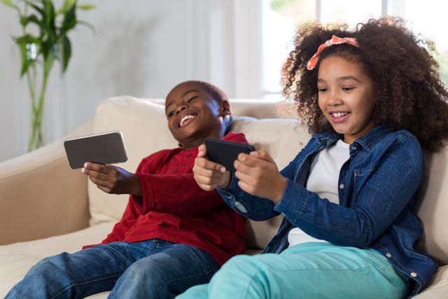 Two children sitting on a couch, smiling and playing games on their smartphones. Ideal for use in articles or advertisements about technology, family activities, or children's entertainment. Perfect for illustrating the impact of technology on modern childhood.