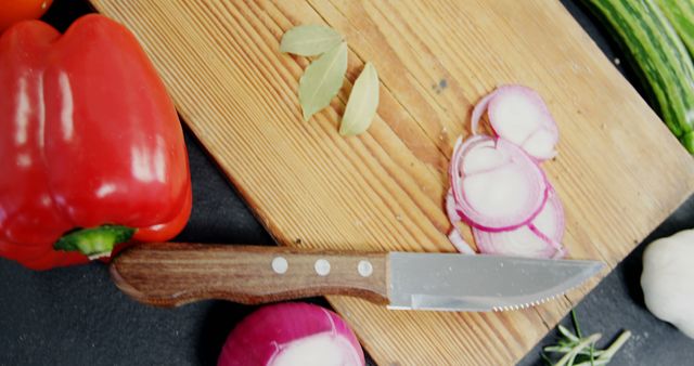 Fresh vegetables including red bell pepper, onion slices, and bay leaves on a wooden cutting board with a kitchen knife. Perfect for use in articles or advertisements about healthy recipes, meal preparation, cooking classes, or kitchen essentials.