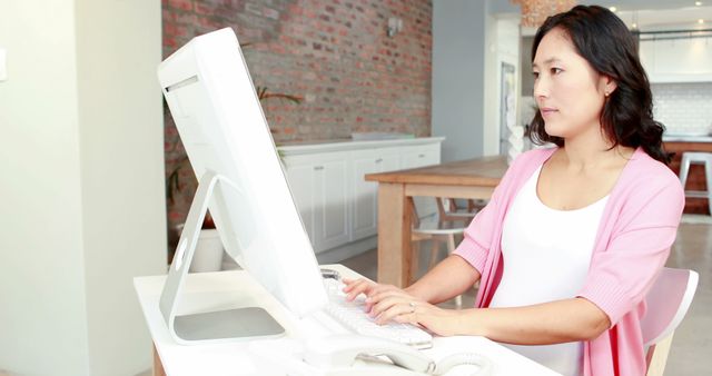 Businesswoman in a casual pink cardigan working at a computer in a modern office space with a white color scheme and exposed brick walls. Suitable for use in business, office work, remote working themes, and articles about work-life balance or home office setups.