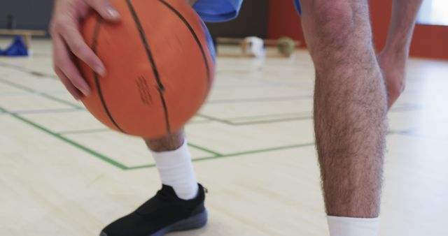 This close-up depicts a male basketball player vigorously dribbling the ball on an indoor court. Ideal for articles, advertising, and sports promotions focusing on basketball training, athletic movements, or sportswear. Suitable for use in sports blogs, fitness magazines, or advertisements promoting basketball gear and sports facilities.