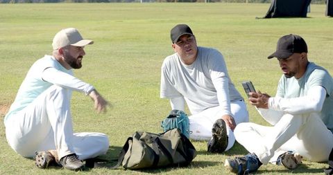 Baseball teammates sitting on grass checking phone reviewing plays and sharing camaraderie