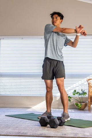 Asian man stretching arms before workout on mat with dumbbells in bright home interior
