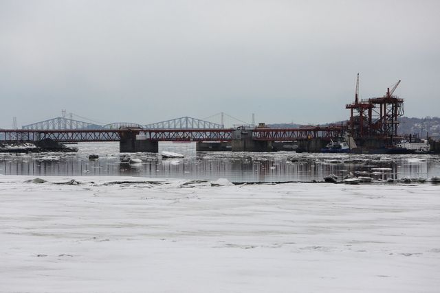 Bridge construction site over a frozen river in winter with cranes and scaffolding. Suitable for illustrating engineering projects, urban development, infrastructure enhancements, and cold weather work settings.