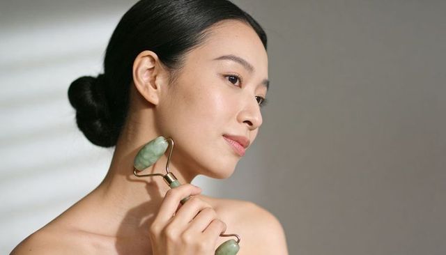 Asian woman rolling jade facial roller along jawline for radiant skincare glow