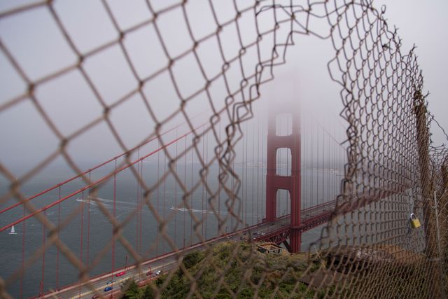 Foggy Golden Gate Bridge viewed through rusted chain link fence. Ideal for travel blogs, urban exploration articles or artwork emphasizing decay or barriers.