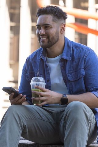 Smiling man drinking green smoothie while checking smartphone on urban steps