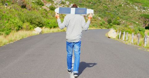 Young man walking down rural road carrying surfboard