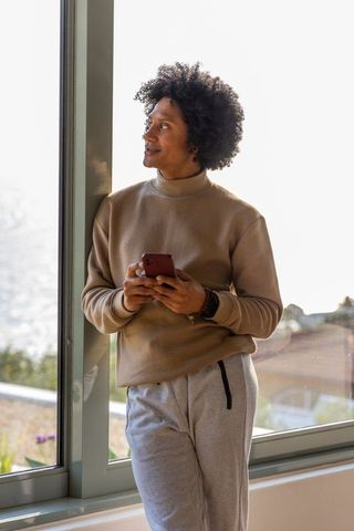 African american man leaning by window holding smartphone contemplating in sunlit interior
