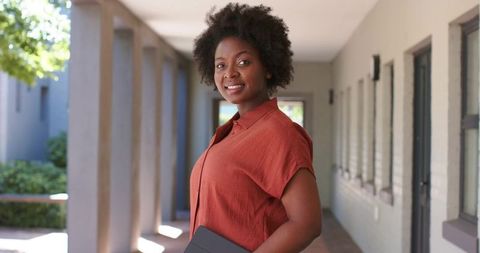 African woman smiling on campus corridor holding laptop folio, wearing rust shirt