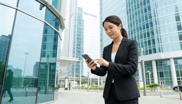 Asian businesswoman checking smartphone outside glass office in financial district