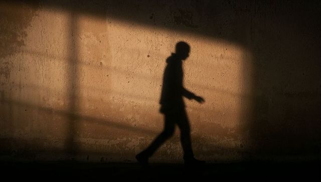 Walking hooded figure casting long shadow on textured concrete wall with rectangular light