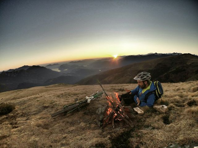 Cyclist is relaxing by campfire on mountain peak during a stunning sunset. Bicycles are resting on ground beside him. Scene captures sense of adventure, tranquility, and connection with nature. Ideal use for outdoor adventure promotion, travel blogs or advertisements, camping gear marketing, seasonal imagery.