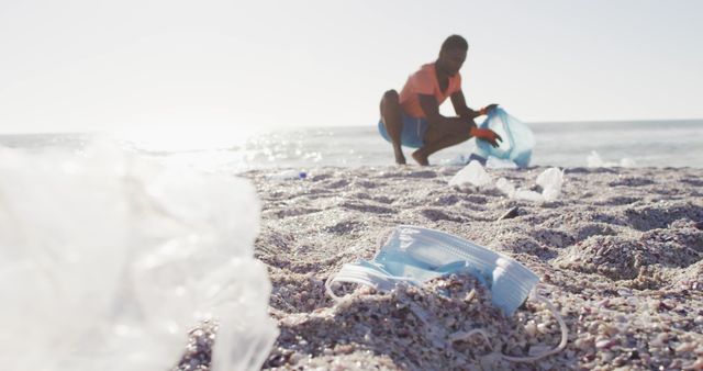 Man wearing casual clothes collecting plastic litter on beach. Foreground focuses on a discarded mask. Can be used for themes on environmental conservation, ecological volunteer activities, coastal cleanups, and raising awareness on plastic pollution.