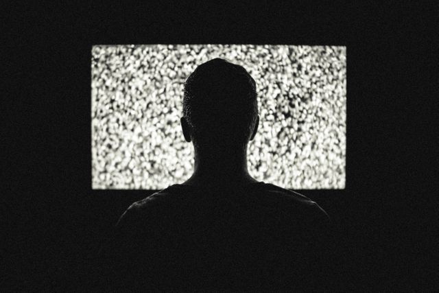 Image showing a person facing an old television screen displaying static in a dark room. Ideal for depicting concepts of media overload, retro technology, eerie ambiance, digital noise, or nostalgic reflections on past technology.