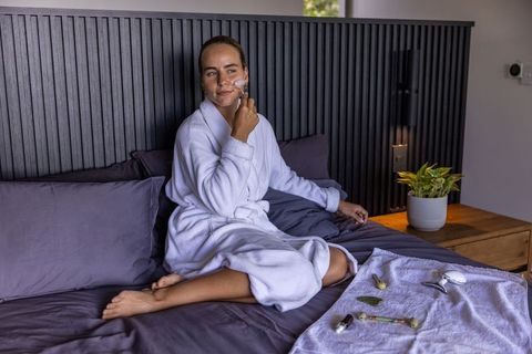 Woman using jade facial roller while relaxing in white bathrobe on bed for morning self care