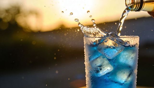 Blue cocktail pouring into frosted glass with sugared rim and ice at golden hour