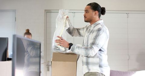 Man unpacking bubble-wrapped gadget from box in modern office, colleagues watching nearby