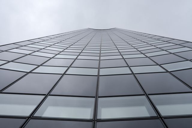 Image of a modern skyscraper captured from a low angle, showcasing its glass facade and symmetrical design. The grey, cloudy sky creates a moody atmosphere. Ideal for use in articles about urban development, corporate landscapes, modern architecture, city infrastructure, or real estate investments.