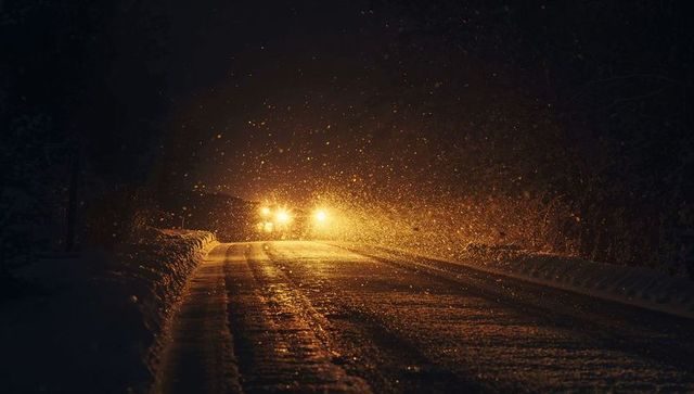 Car headlights piercing heavy snowfall on dark rural road with glossy tire tracks