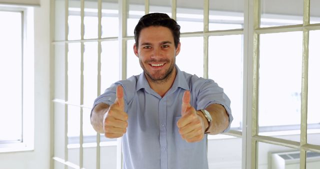 A cheerful man in business attire standing in an office, giving a thumbs up gesture, conveying success and approval. Perfect for use in corporate promotions, motivational content, advertisements, team-building materials, or any context requiring a positive and confident image.