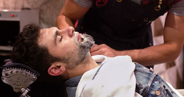 Man receiving a professional shave from a barber. Perfect for illustrating barber services, male grooming routines, and self-care practices. Suitable for use in advertising barber shops, grooming products, or magazine articles on men's personal care.
