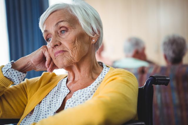 Senior woman sitting in a wheelchair looking worried, with hand holding her head. Ideal for use in articles or advertisements related to elderly care, nursing homes, mental health, aging, and healthcare services. Can also be used in brochures or websites focusing on senior living and retirement communities.