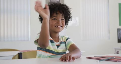 Smiling schoolboy raising hand in classroom at desk with notebooks and pencil engaged